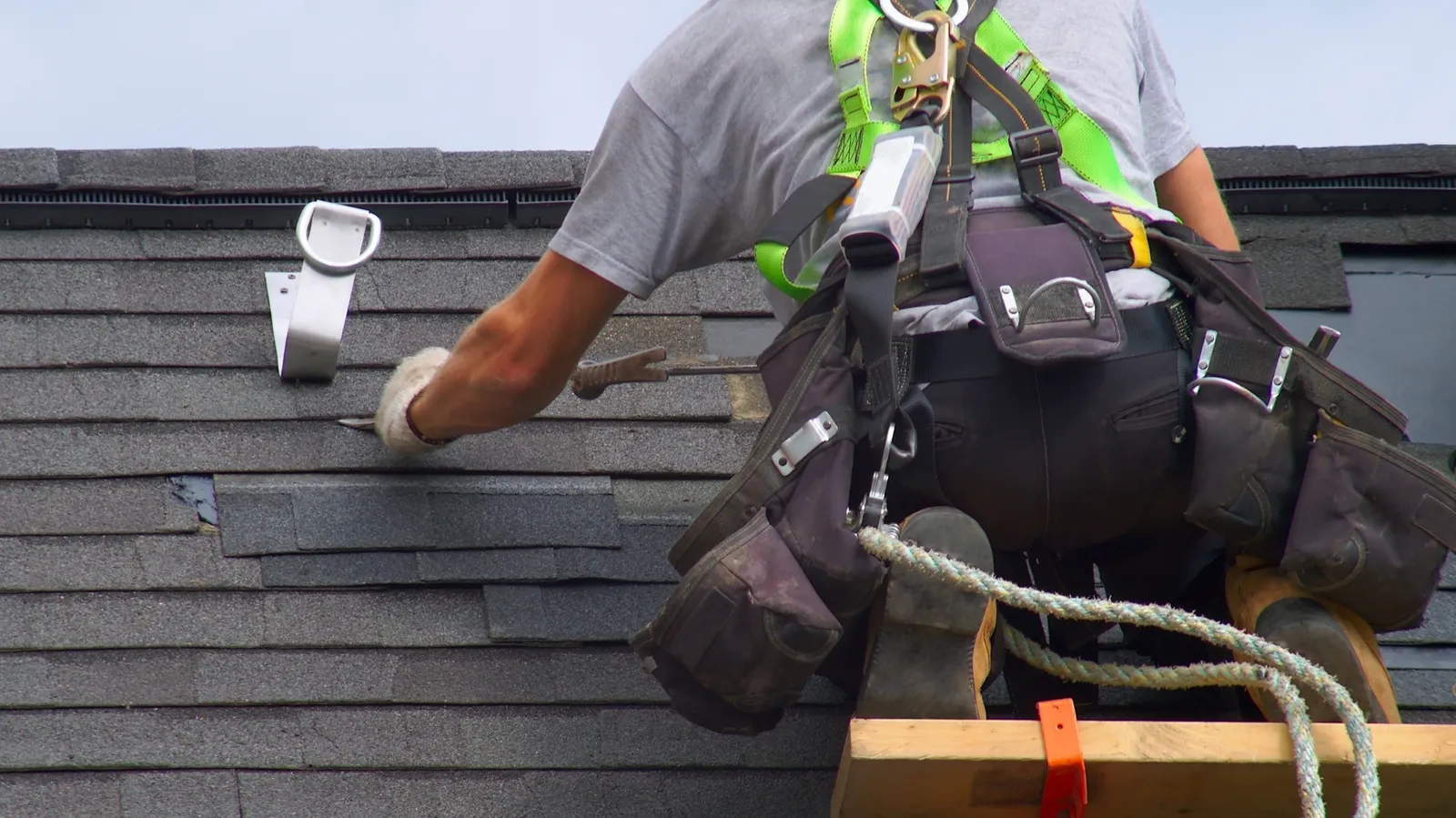 On a gray roof a roofer wearing a safety harness fits a shingle