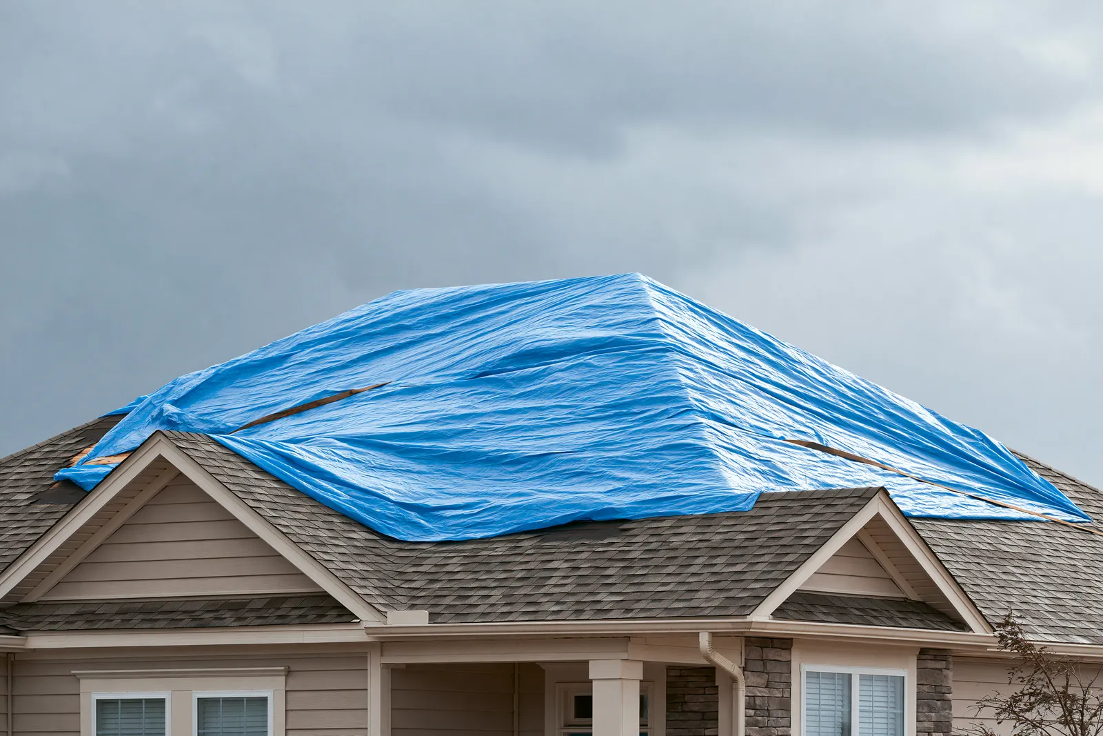 Under grey clouds a blue tarp shields storm-damaged roof sections on a two-story home
